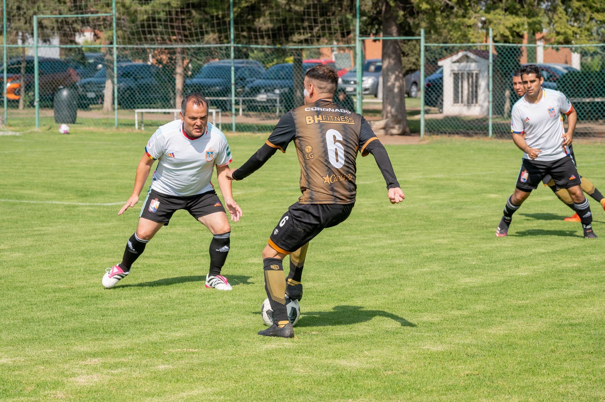 Adults playing a Sunday-league soccer match on an ordinary grass pitch, cars and a chain-link fence behind them